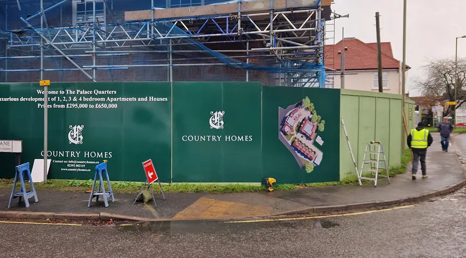 How much does Hoarding Signage Cost 4 Workers installing large green "Country Homes" branded hoarding panels around a housing development site with scaffolding in the background.