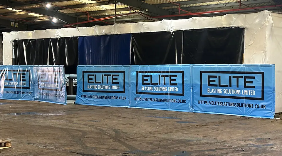 heras-acoustic-barriers-elite-blasting-solutions-1 Five light blue custom printed acoustic barriers with the Elite Blasting Solutions logo set up inside a large industrial warehouse, shielding a white and black enclosed grit blasting structure.