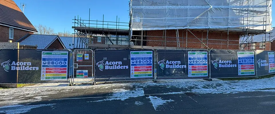 Heras Fence Covers for Acorn Builders 2 A wide-angle view of a residential construction site enclosed by a continuous row of black Heras fence covers. The covers feature repeating Acorn Builders logos and site safety signs.