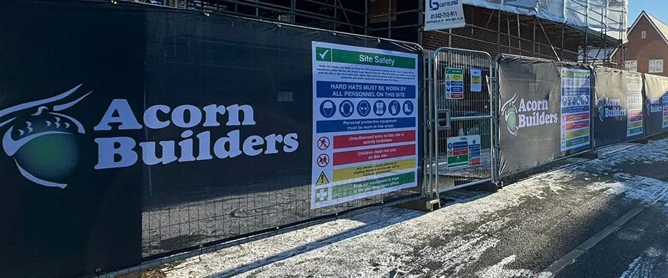 Heras Fence Covers for Acorn Builders 3 A close-up view of a black printed Airmesh Heras fence cover with white and green "Acorn Builders" logo on the left and a large, brightly colored "Site Safety" compliance sign on the right.