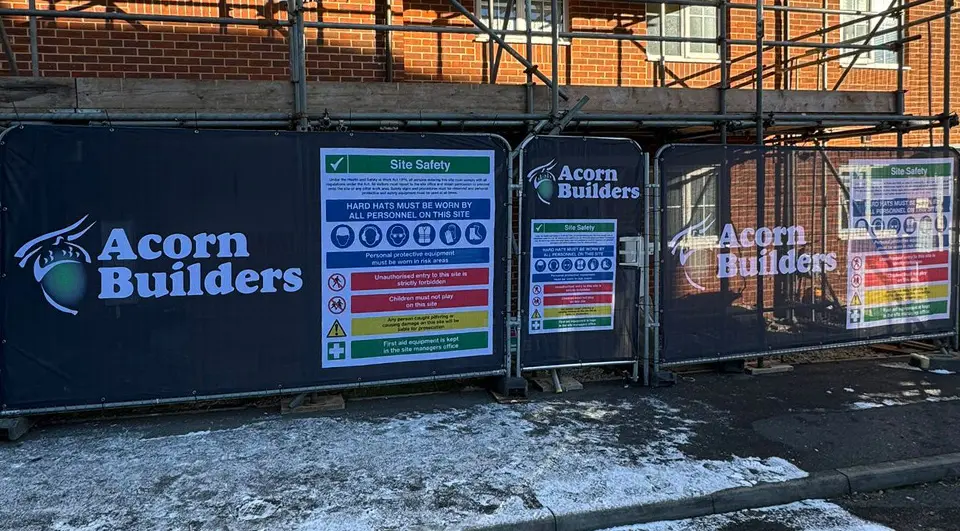 heras-fence-covers-acorn-builders Black Airmesh fence covers and a matching pedestrian gate cover prominently displaying the Acorn Builders branding and site safety regulations.