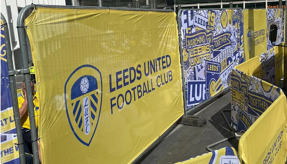 Leeds United FA Cup Semi-Final Branding at Wembley Stadium 2 A close-up view of a tall, mesh Heras fence cover featuring the Leeds United Football Club crest on a bold yellow background next to an intricate blue and yellow collage design.