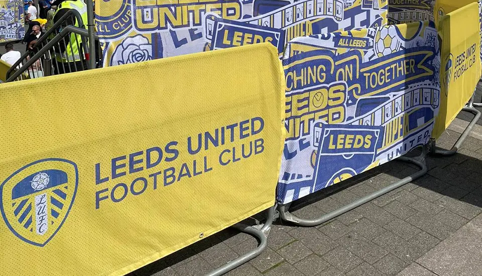 Leeds United FA Cup Semi-Final Branding at Wembley Stadium 4 Low-profile metal MET crowd control barriers lined up on a paved walkway, fitted with custom yellow and blue Leeds United airmesh covers.