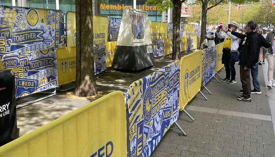 leeds-united-at-wembley-case-study Fans standing outside Wembley Library taking photos in front of metal crowd control barriers dressed in vibrant yellow and blue Leeds United branded covers.