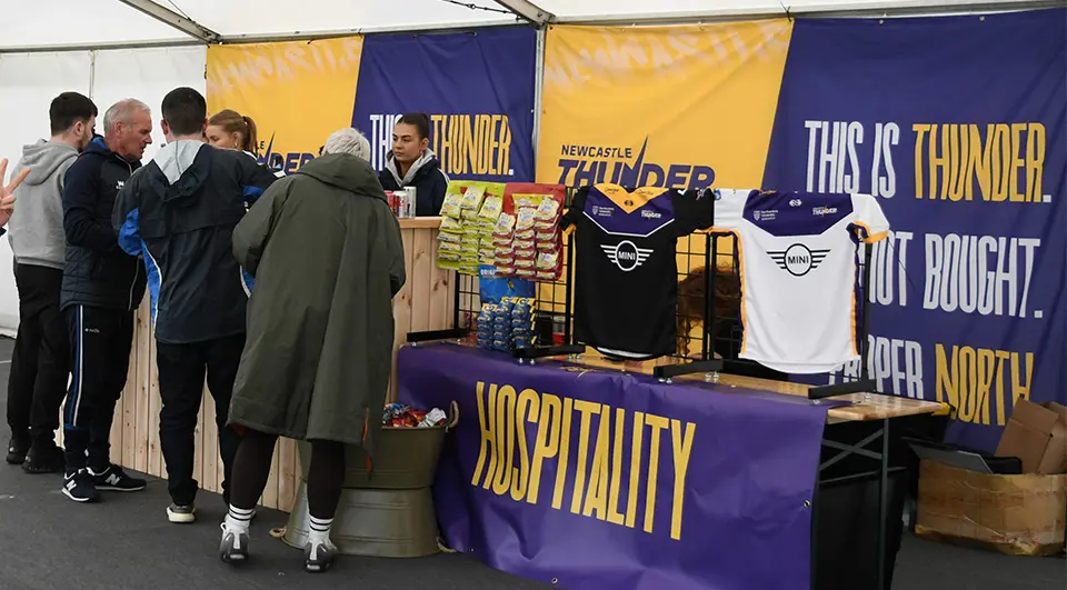 newcastle-thunder-custom-banners Fans gather around a Newcastle Thunder merchandise and hospitality stall inside a white marquee tent. The stall is dressed with a purple banner reading "HOSPITALITY" in large yellow font, and the background features large blue and yellow custom banners with the slogans "THIS IS THUNDER" and "PROPER NORTH."