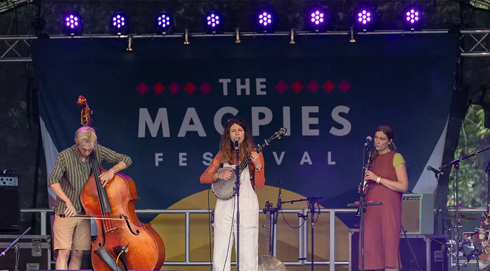 The Magpies Festival stage backdrop with festival branding behind a folk trio performing