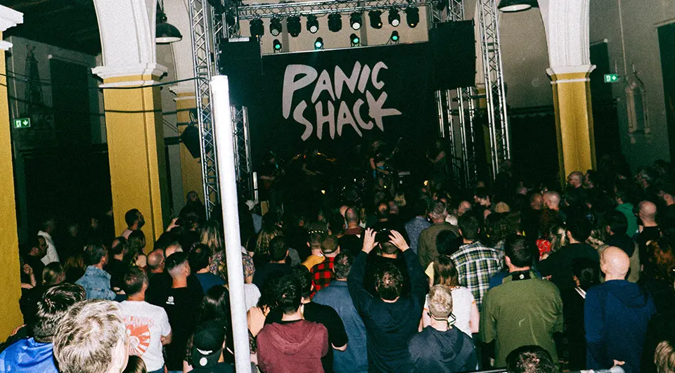 Panic Shack band backdrop with bold white hand-drawn logo on black, above a packed crowd