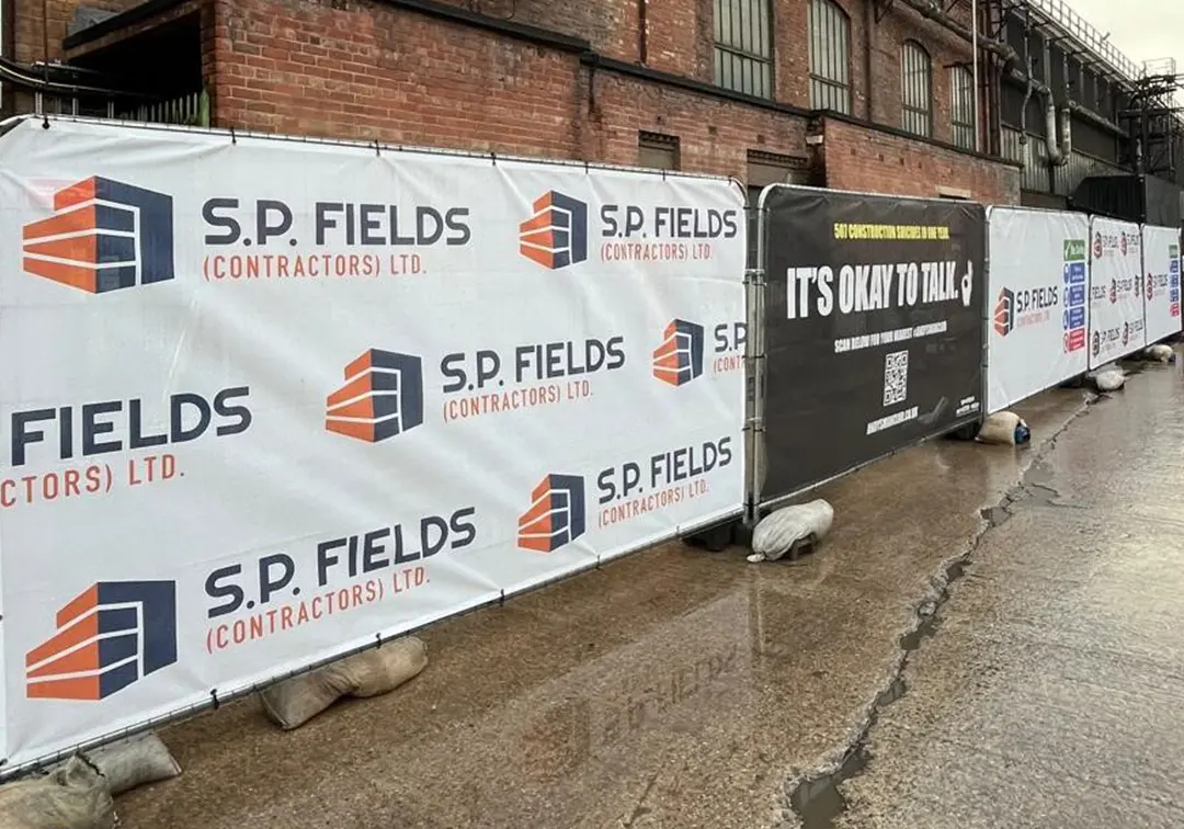 heras-fence-covers-case-study-sp-fields-category-tile Long run of white PVC Mesh Heras fence covers with S.P. Fields (Contractors) Ltd branding, with a black Andy’s Man Club cover in the line, outside a red-brick industrial building.
